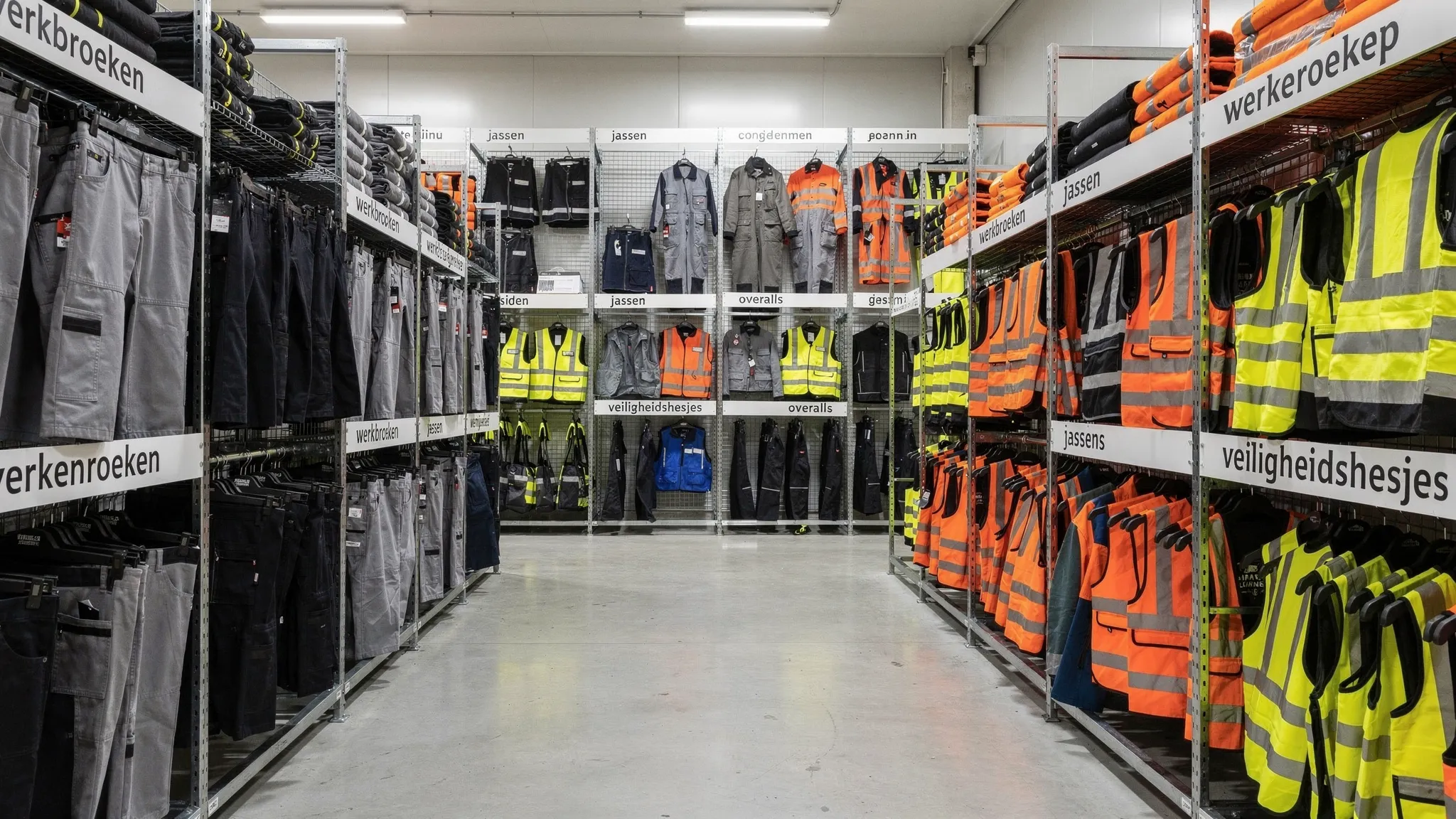 Een overzichtelijke voorraadruimte met professionele werkkleding, waaronder werkbroeken, jassen, overalls en veiligheidshesjes, netjes gesorteerd op rekken en planken voor snelle uitlevering aan dealers en zakelijke klanten.