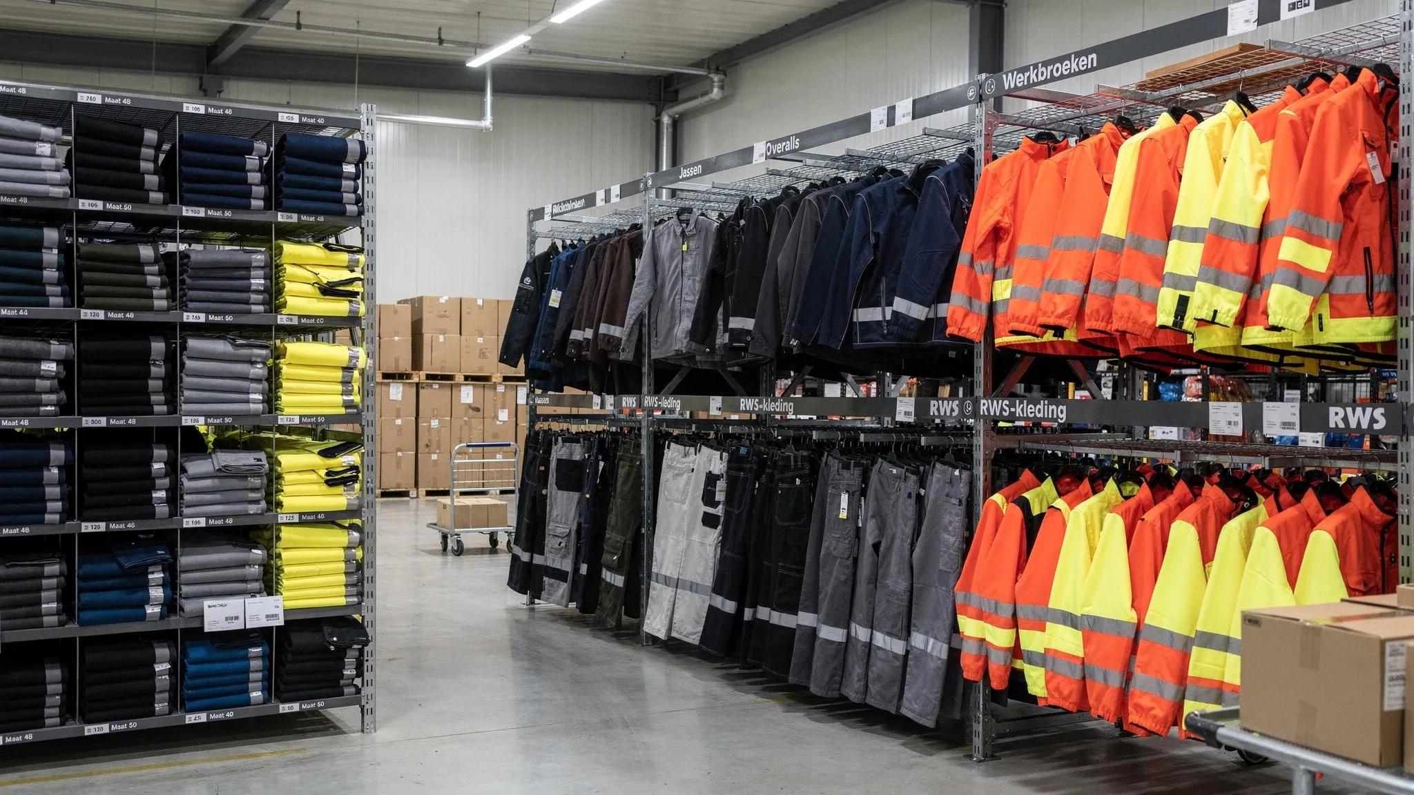 Magazijnstellingen met verschillende soorten professionele werkkleding, waaronder werkbroeken, jassen, overalls en RWS-kleding, overzichtelijk gesorteerd op maat en kleur voor snelle levering aan resellers en zakelijke klanten.
